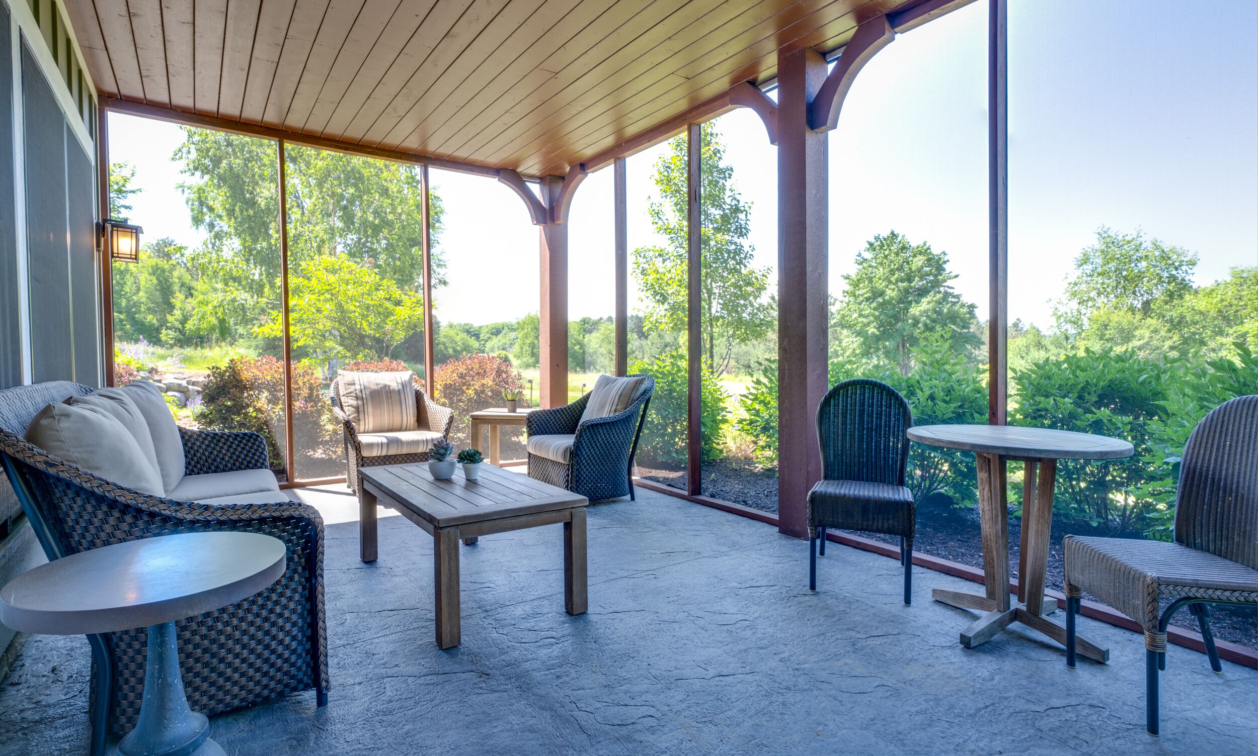 Ein überdachter Patio-Bereich mit Korbmöbeln, einem Holztisch und Blick auf einen üppigen Garten.