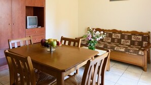 A dining area with a wooden table, chairs, and a fruit bowl. There is a wooden cabinet with a television and a sofa with a patterned cushion. - Argyro Apartments (Agios Nikolaos)