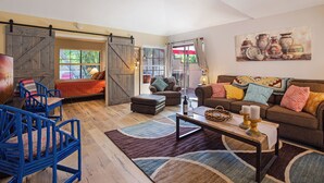 A spacious living room with a large sofa, a coffee table, and blue chairs. There is a sliding barn door and a painting on the wall. - Oasis with 3rd Bedroom/Den, sleeps 6, 3 patios, adult and childrens bikes (Palm Springs)
