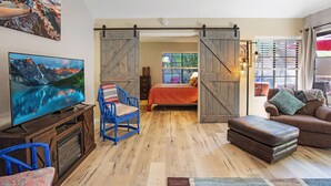 A room with a wooden floor, a bed with a red blanket, a television, a blue chair, and a brown couch. - Oasis with 3rd Bedroom/Den, sleeps 6, 3 patios, adult and childrens bikes (Palm Springs)