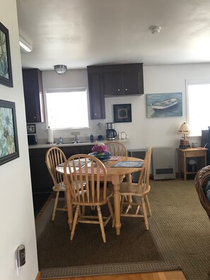 A dining area with a wooden table and chairs, a kitchen with dark cabinets, a window, and a painting of a boat. - The Boathouse (Eureka)