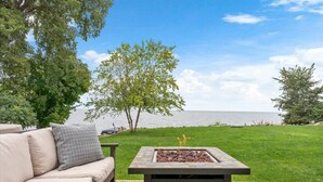 Eine Außenterrasse mit einem Steinkamin, einem beigen Sofa mit grauem Kissen und Blick auf den See.