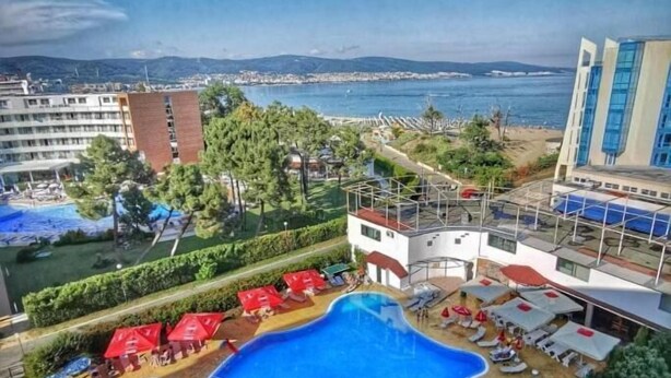 Una piscina de forma singular, rodeada de sillones de descanso y sombrillas, con vista al mar y edificios al fondo.