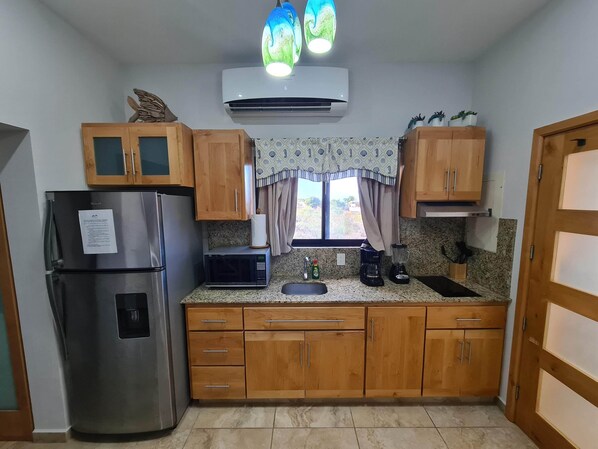 Cocina compacta con armarios de madera, un refrigerador de acero inoxidable, un microondas, una cafetera y una ventana con cortinas.