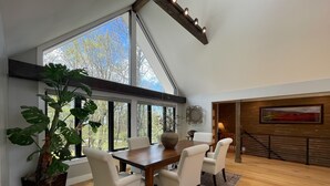 Un coin repas avec une table en bois, des chaises blanches et une grande fenêtre donnant sur les arbres.