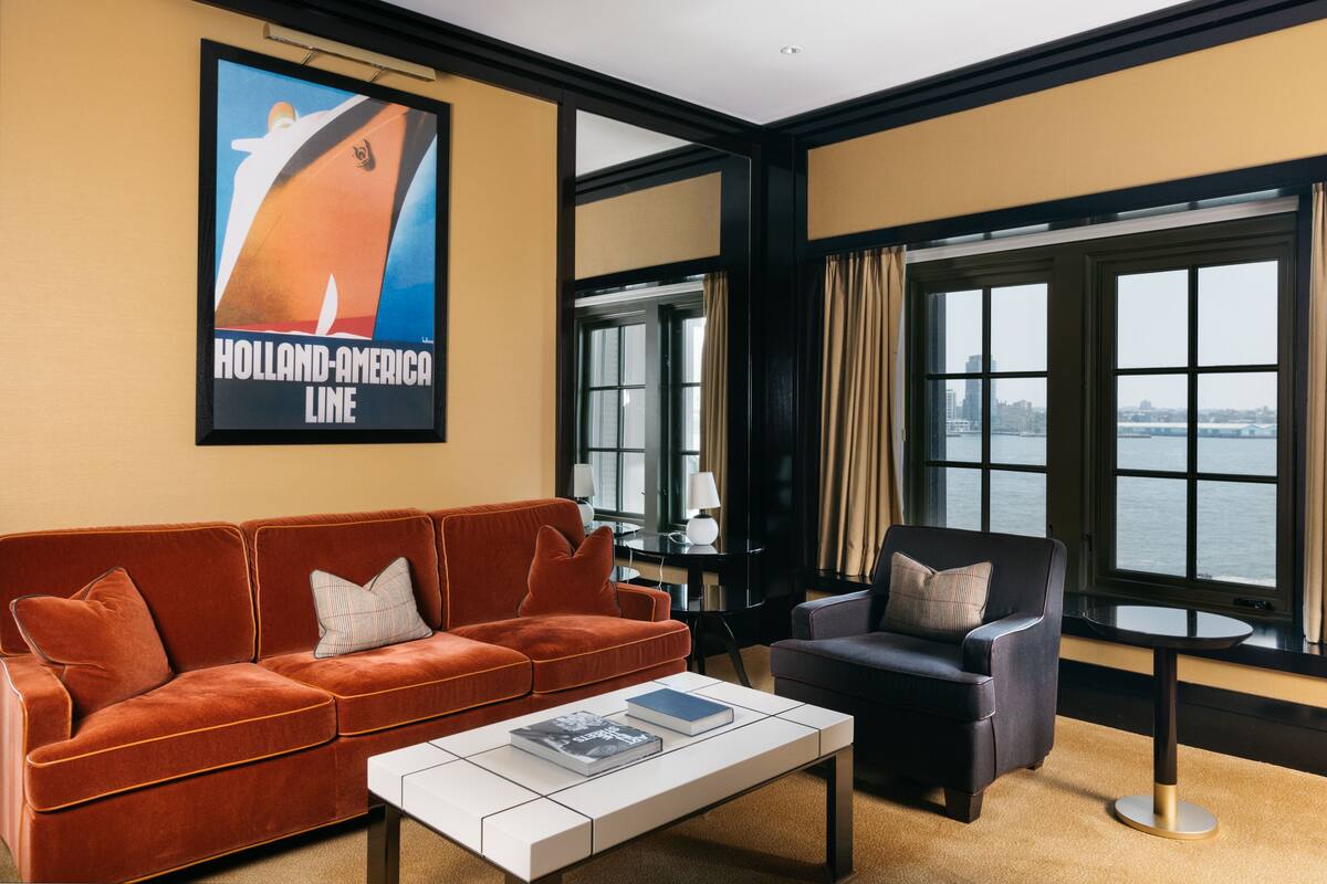 Un salon moderne avec un canapé, un fauteuil et une table basse, une affiche représentant un navire et de grandes fenêtres donnant sur l’eau.