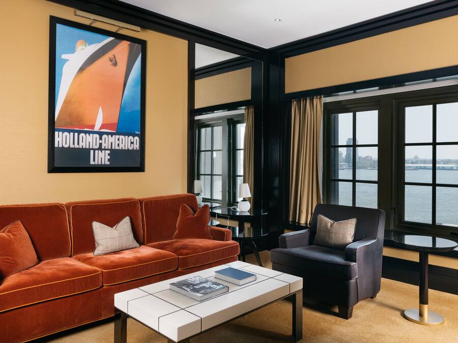 Een moderne woonkamer met een bank, fauteuil en salontafel, een poster van een schip, en grote ramen met uitzicht op het water.