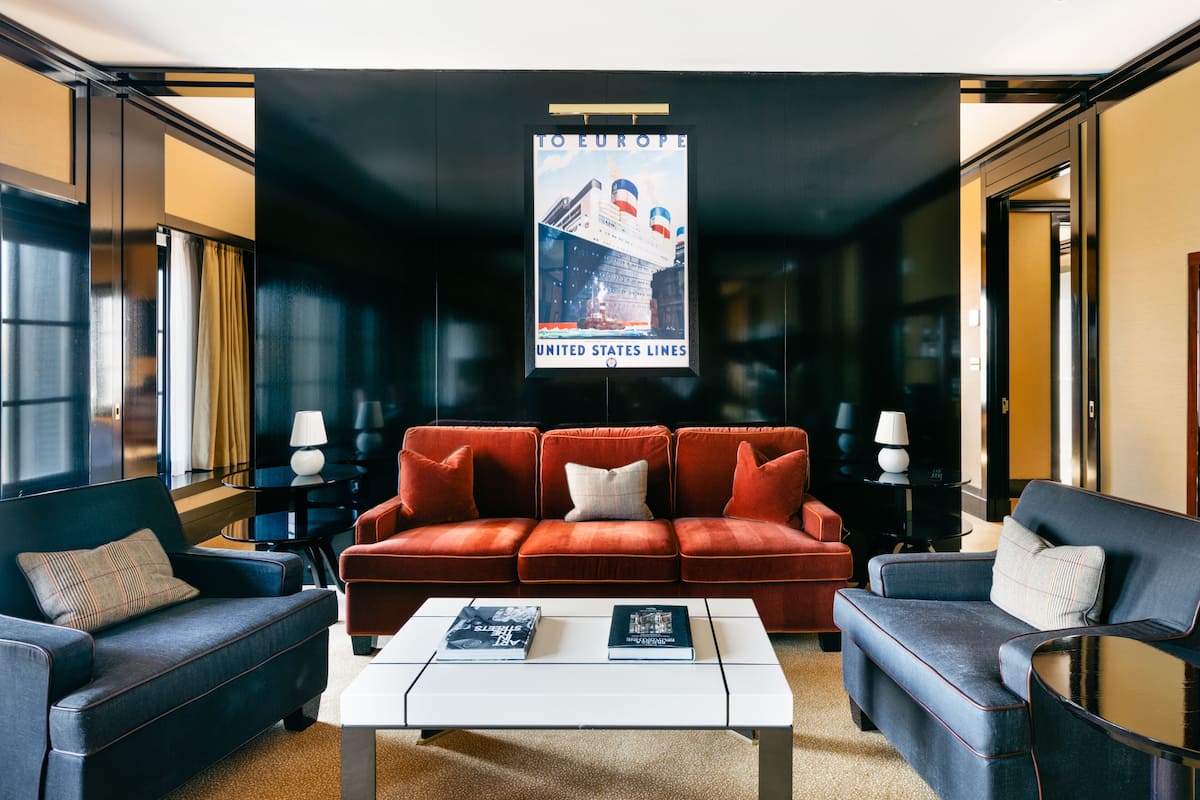 Un hall d’hôtel moderne avec un canapé rouge, deux fauteuils, une table basse et une affiche représentant un navire de la compagnie United States Lines.