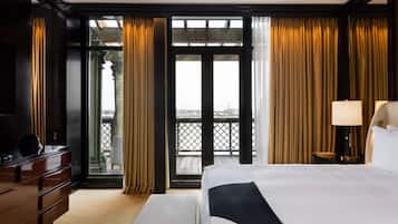 A hotel room with a large bed, a dresser, a lamp, and a view of the city through a balcony.