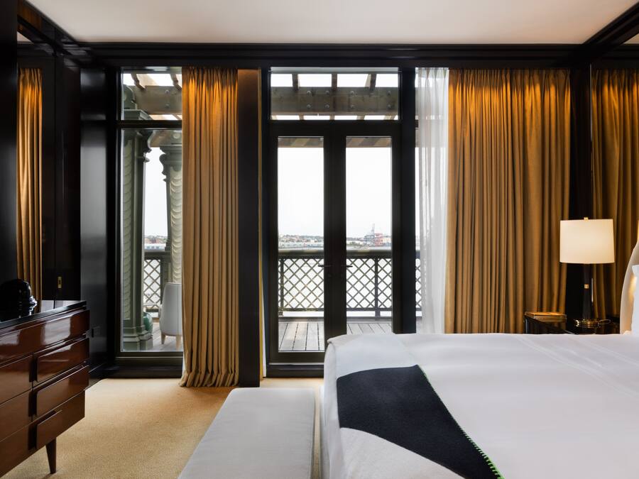 A hotel room with a large bed, a dresser, a lamp, and a view of the city through a balcony.