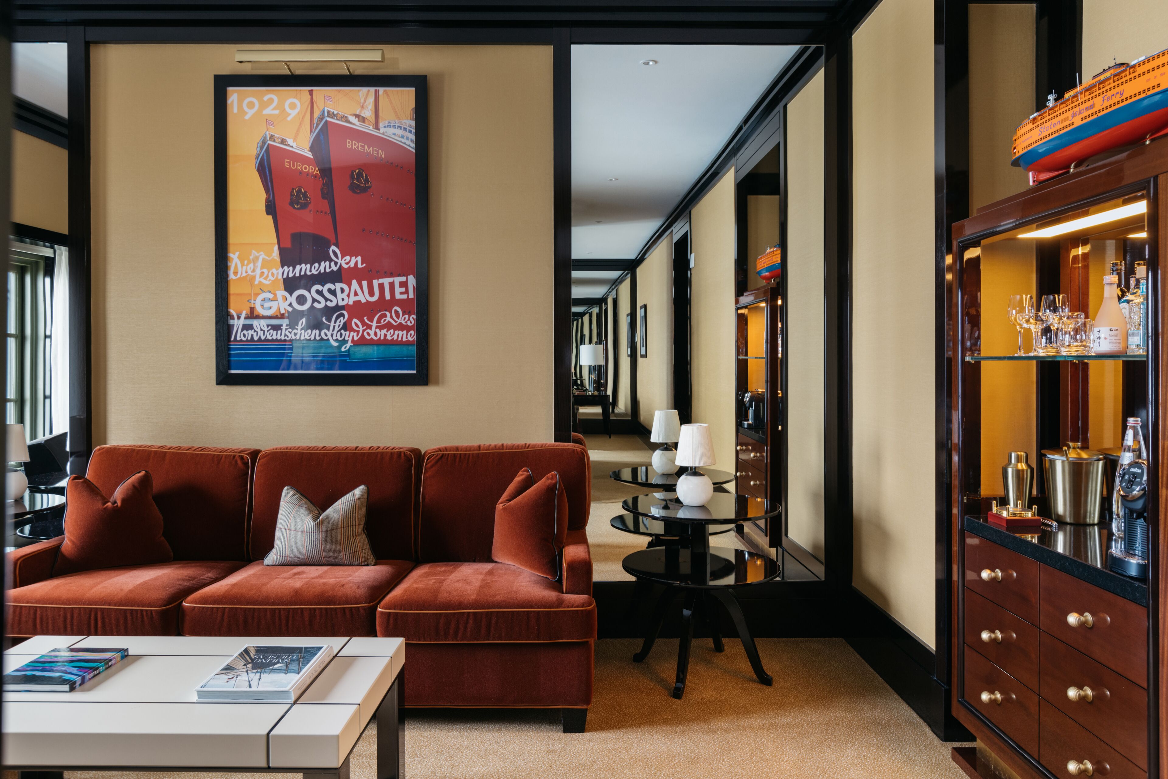 A hotel lobby with a red sofa, a coffee table, a poster, a round table with lamps, and a cabinet with bottles.