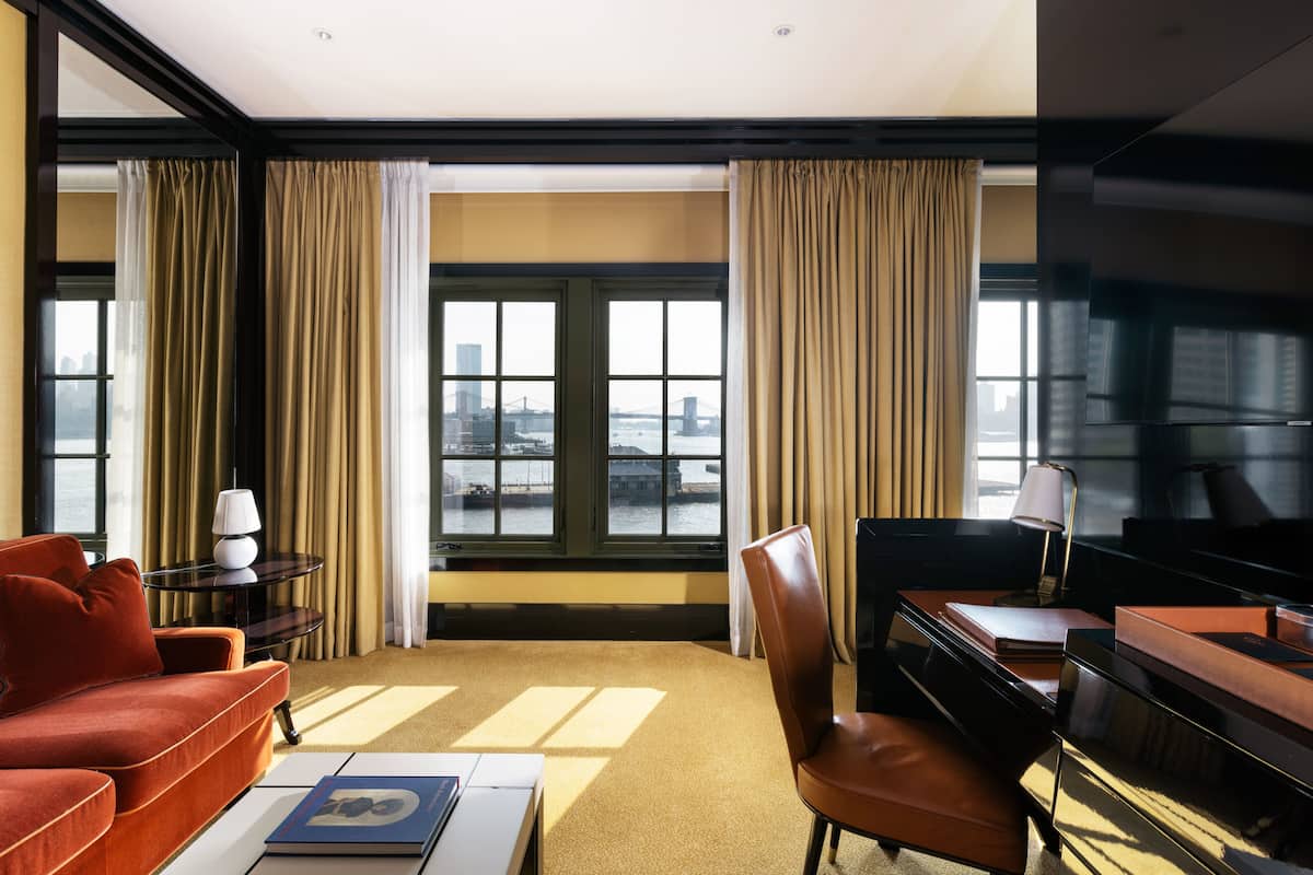 Une chambre d’hôtel avec vue sur la ville, un canapé rouge, un fauteuil en cuir marron, un bureau et un piano.