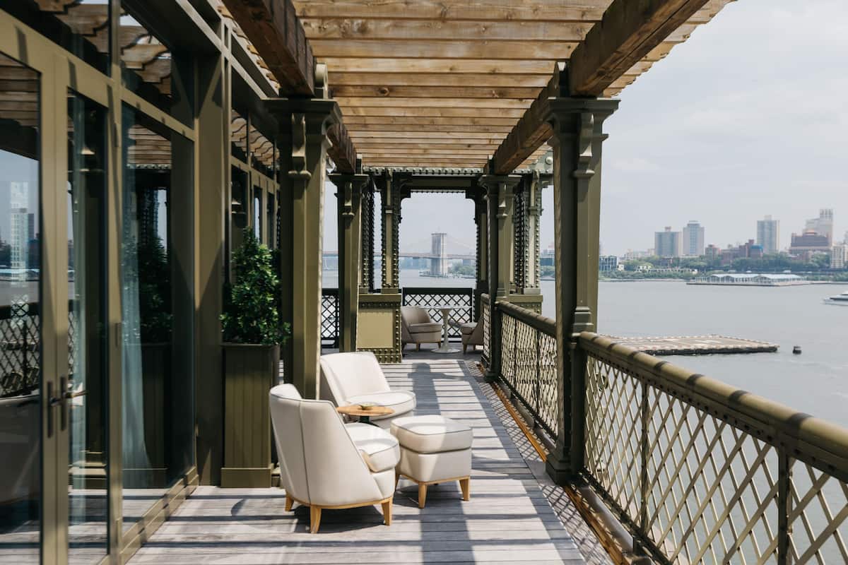 Un espace extérieur aménagé avec une zone de repos couverte, comprenant deux chaises blanches rembourrées et une petite table, offrant une vue sur une étendue d’eau et le panorama de la ville.