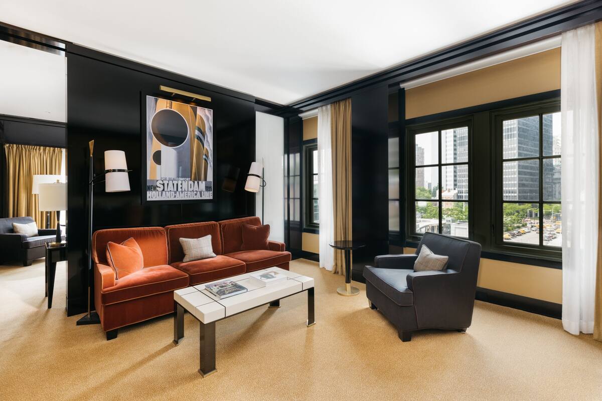 Un salon moderne avec un canapé rouge, un fauteuil gris et une table basse. Une grande fenêtre offre une vue sur les immeubles, et une affiche est accrochée au mur.