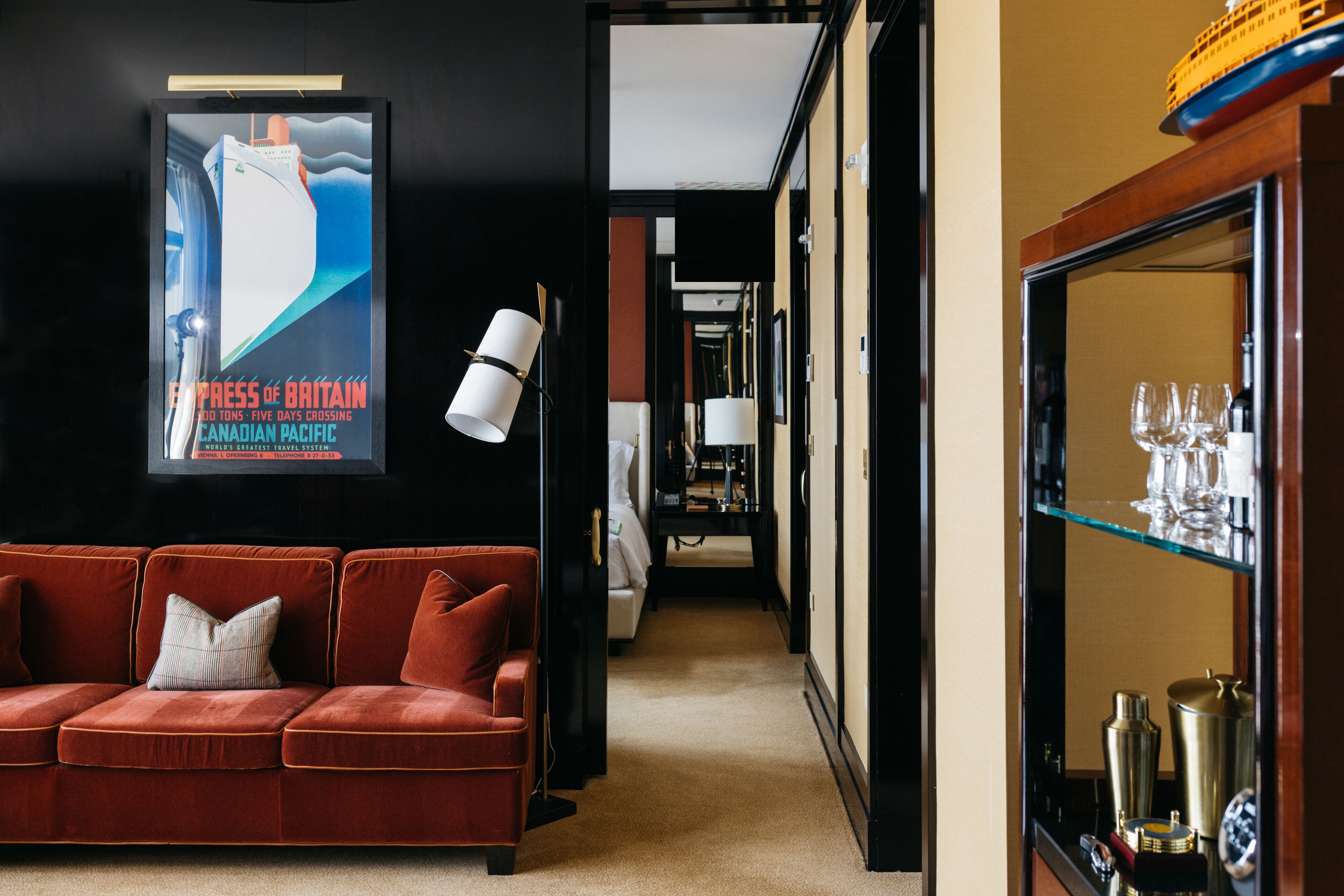 A hotel lobby with a red sofa, a poster of a ship, and a glass display case with bottles.
