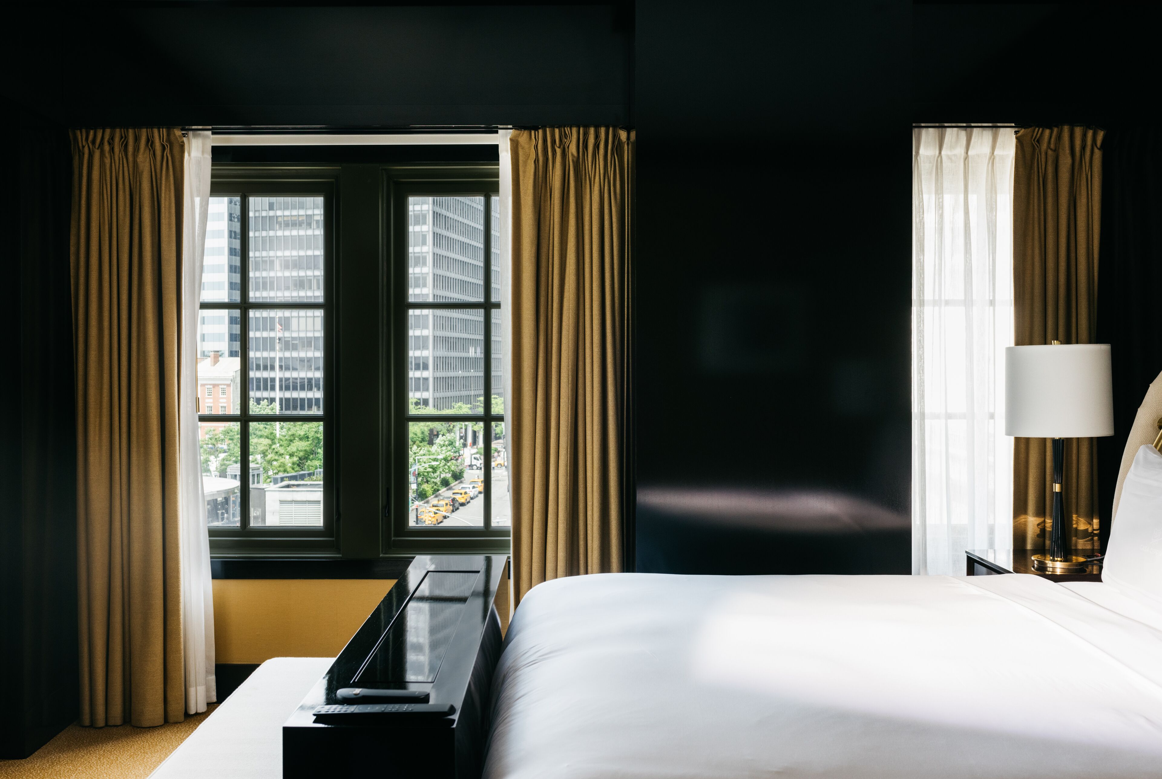 Ein Hotelzimmer mit Bett, Schreibtisch, Lampe und einem fenster mit Vorhängen.