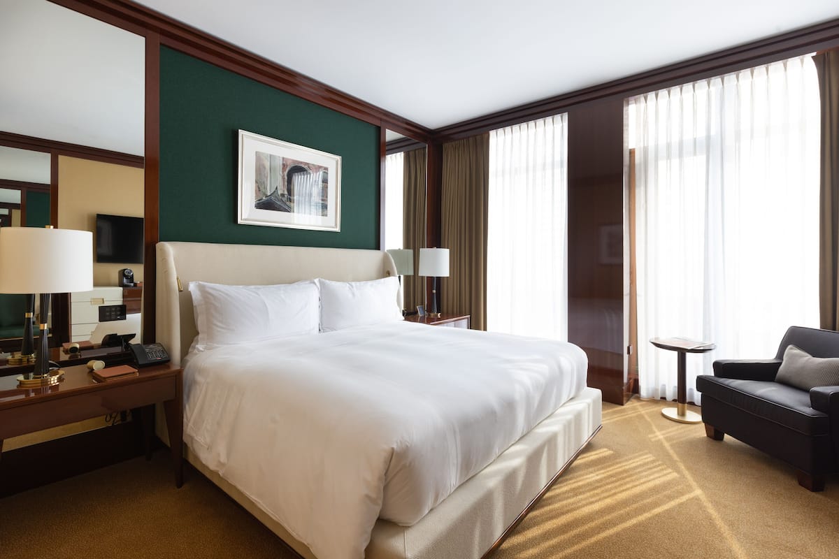 Une chambre d’hôtel avec un grand lit, un bureau avec une lampe, une chaise, une petite table et une grande fenêtre avec des rideaux.
