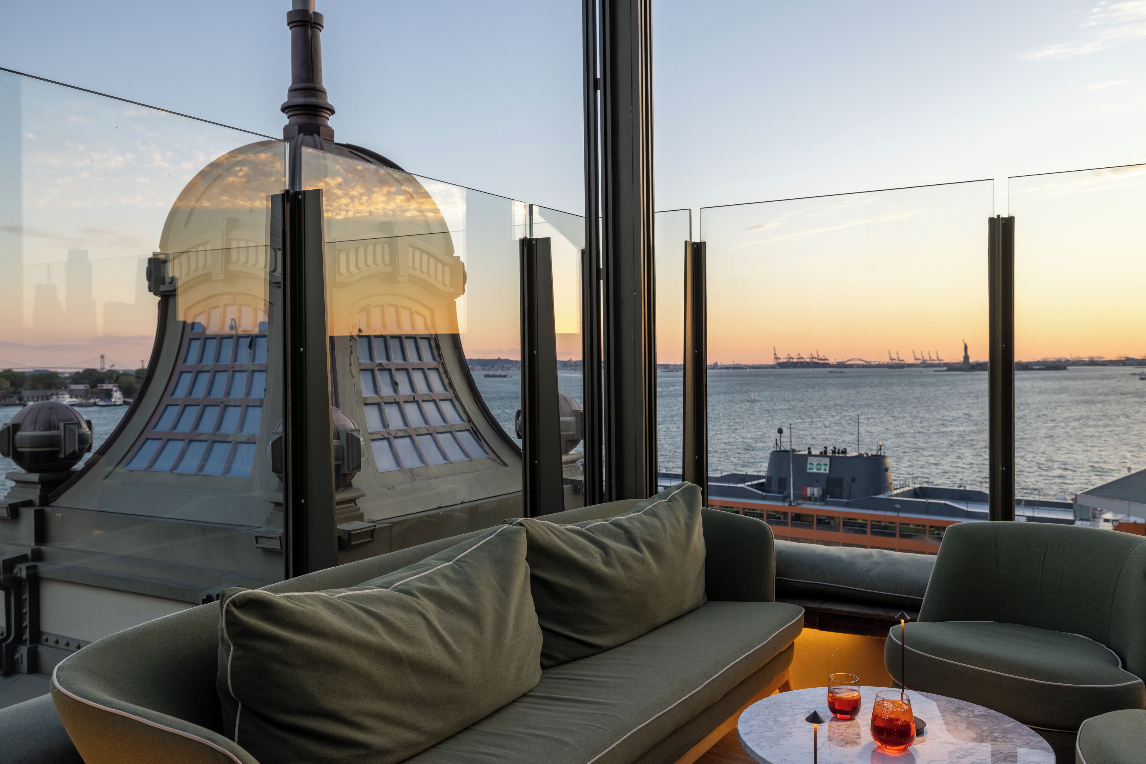 A modern interior with a sofa and a table, overlooking a body of water with a bridge and ships in the distance.