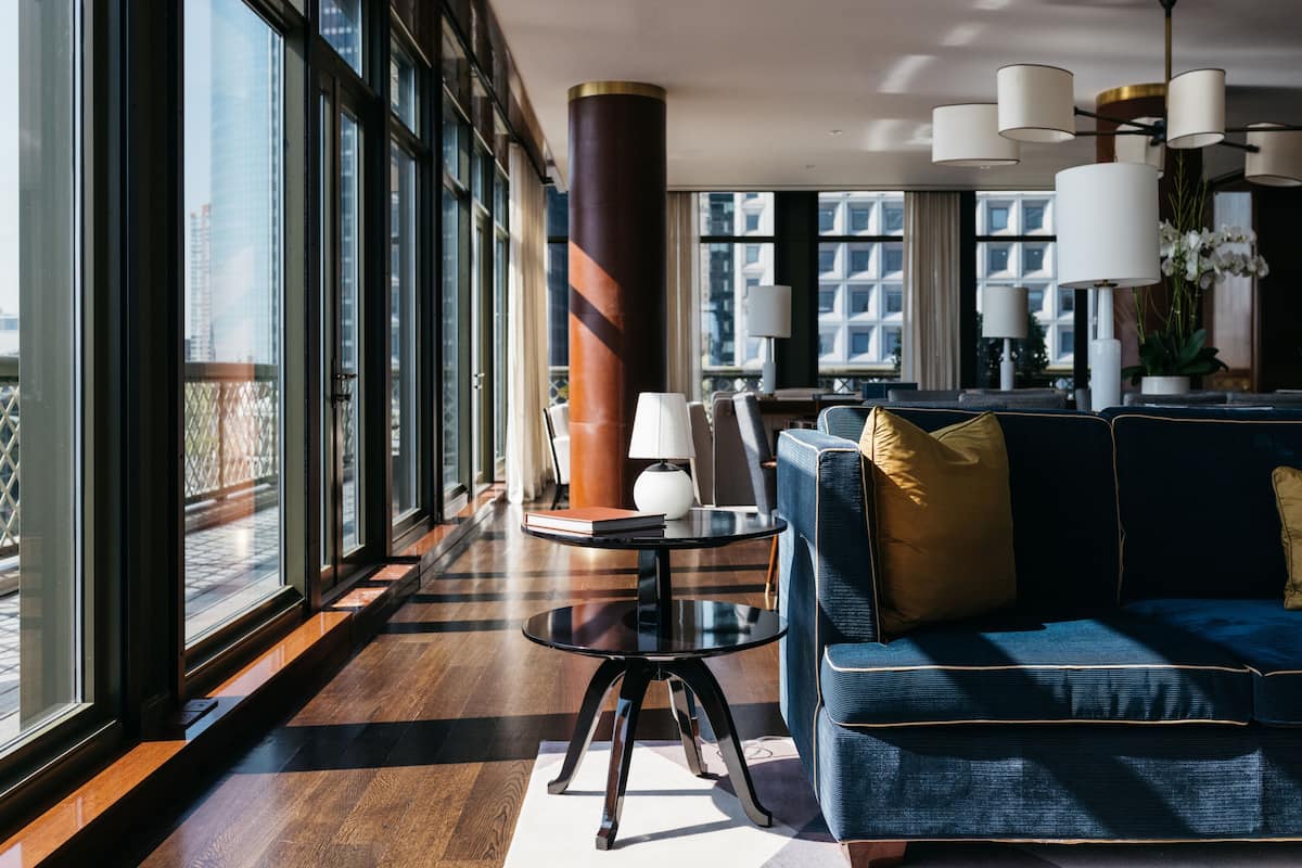 Un hall d’hôtel moderne avec une grande fenêtre, un canapé bleu, une table ronde et plusieurs lampes.