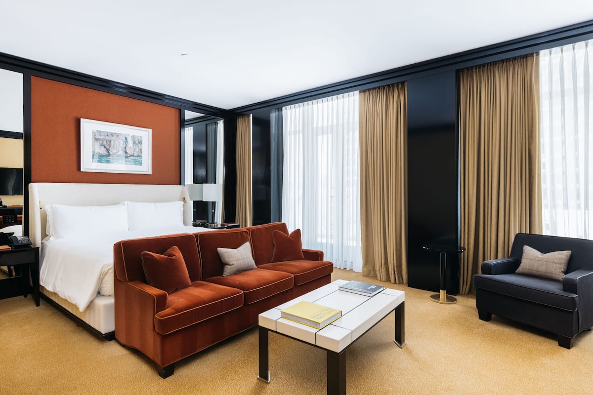 Une chambre d’hôtel avec un lit, un canapé marron, un canapé noir, une table basse blanche et une grande fenêtre avec des rideaux.