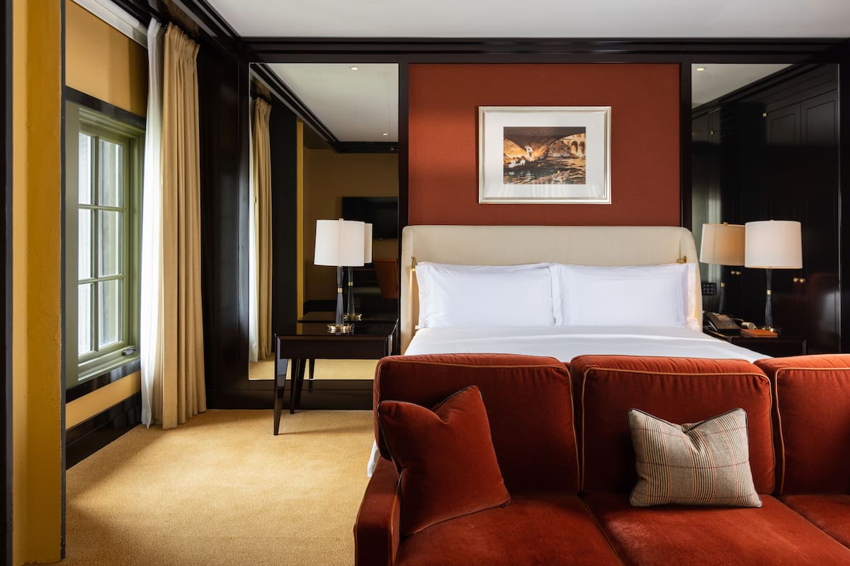 Une chambre d’hôtel avec un grand lit, deux tables de chevet avec des lampes, un canapé rouge avec des coussins et un tableau encadré au mur.