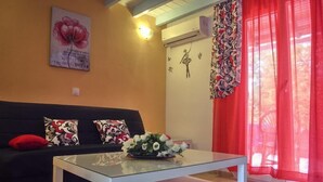 Ein Wohnzimmer mit einer schwarzen Couch, gemusterten Kissen, einem Glastisch mit Blumenarrangement, einer Wand-Klimaanlage und einem Fenster mit roten Vorhängen.