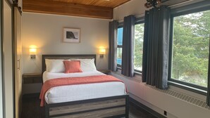 Quarto com cabeceira de madeira, cama com roupa de cama branca e vermelha, duas luminárias de mesa, janela com cortinas e um quadro na parede.