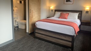Quarto com cama, mesa de cabeceira, quadro na parede e banheiro visível por uma porta aberta.