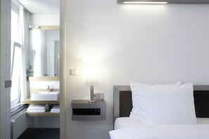 A hotel room with a bed, a nightstand with a lamp, a window with a view of the bathroom, and a wall-mounted shelf with towels. - Beaumont (Maastricht)