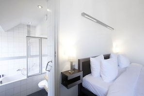 A hotel room with a shower, toilet, bed, bedside table, and a nightstand with a lamp. - Beaumont (Maastricht)