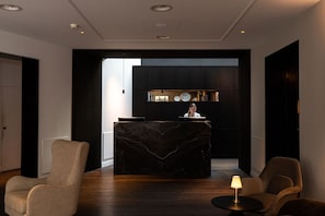 A modern reception area with a marble reception desk, two armchairs, a small table with a lamp, and a bookshelf in the background. - Beaumont (Maastricht)