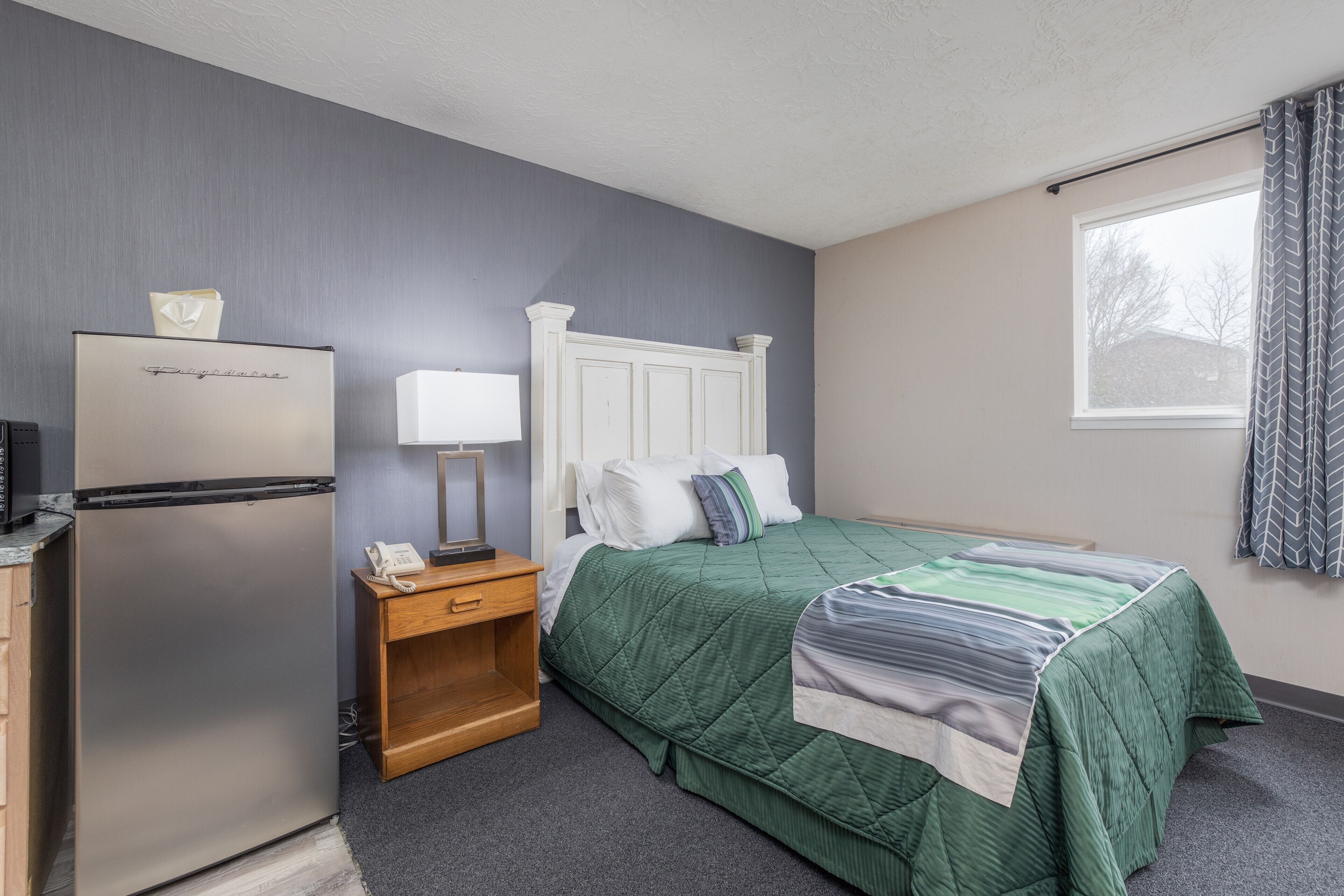 A bedroom with a bed, a nightstand, a refrigerator, and a window with curtains.