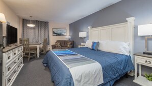 A hotel room with a large bed, a desk, and a chair. There is a dresser with drawers and a television on top.