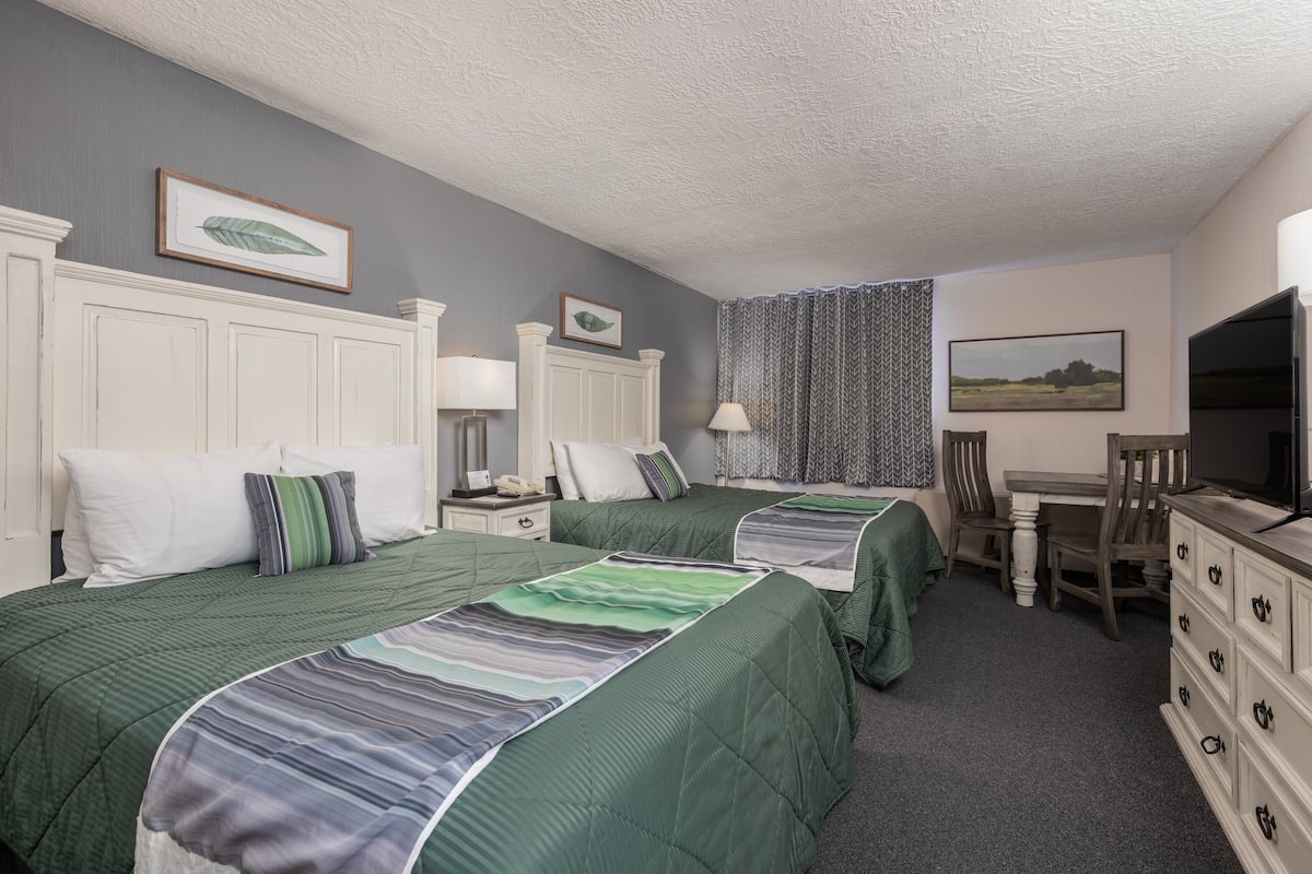 A hotel room with two beds, a desk, a chair, a dresser, and a television.