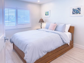 Un dormitorio con cama, ventana con persianas, lámpara de noche y dos cuadros en la pared.