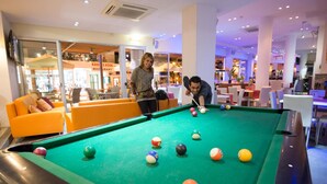 A pool table in a brightly lit indoor area with orange seating and a visible restaurant in the background. - Eligonia Apartments (Ayia Napa)