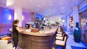 A bar area with a counter, stools, and a bartender. There are shelves with bottles and a television screen. - Eligonia Apartments (Ayia Napa)