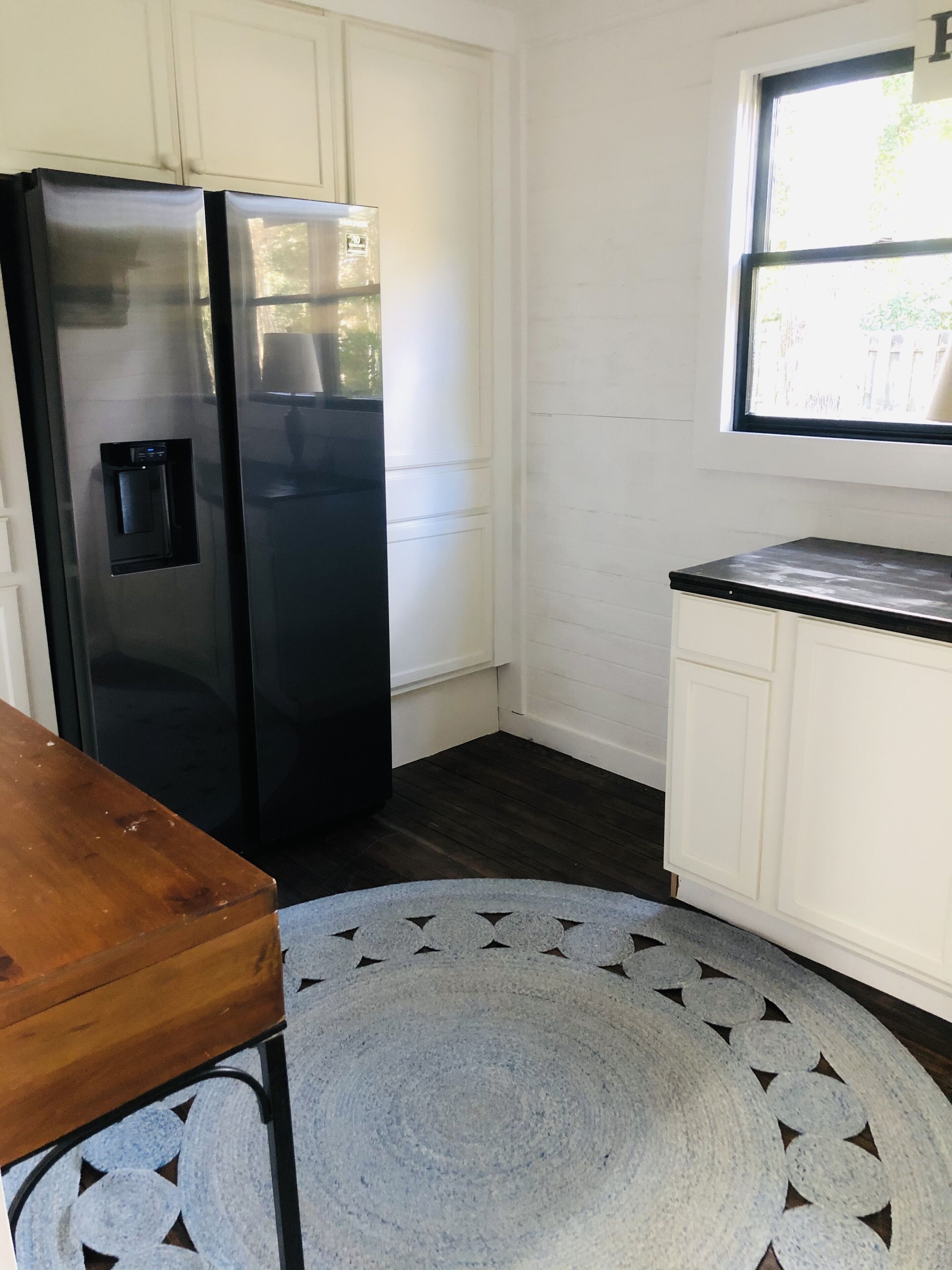 A kitchen with white cabinets, a black refrigerator, a wooden table, and a patterned rug.