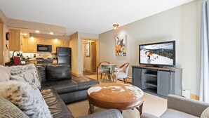 A modern living room with a sofa, ottoman, and television. There is a kitchen area with a microwave and refrigerator.