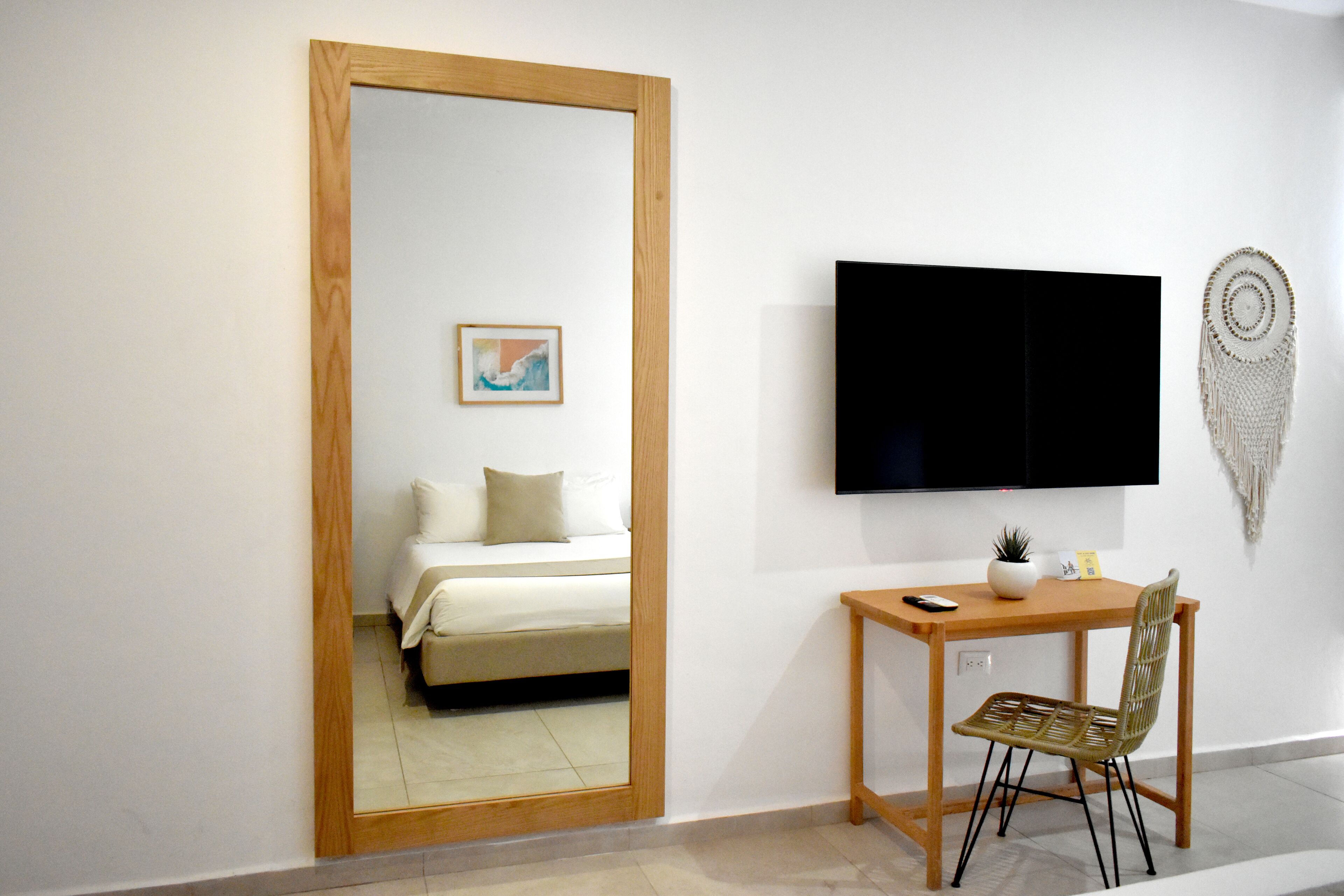 Une chambre d’hôtel avec un miroir à cadre en bois, un lit avec une literie blanche, un petit bureau avec une chaise, une télévision et un élément décoratif fixé au mur.