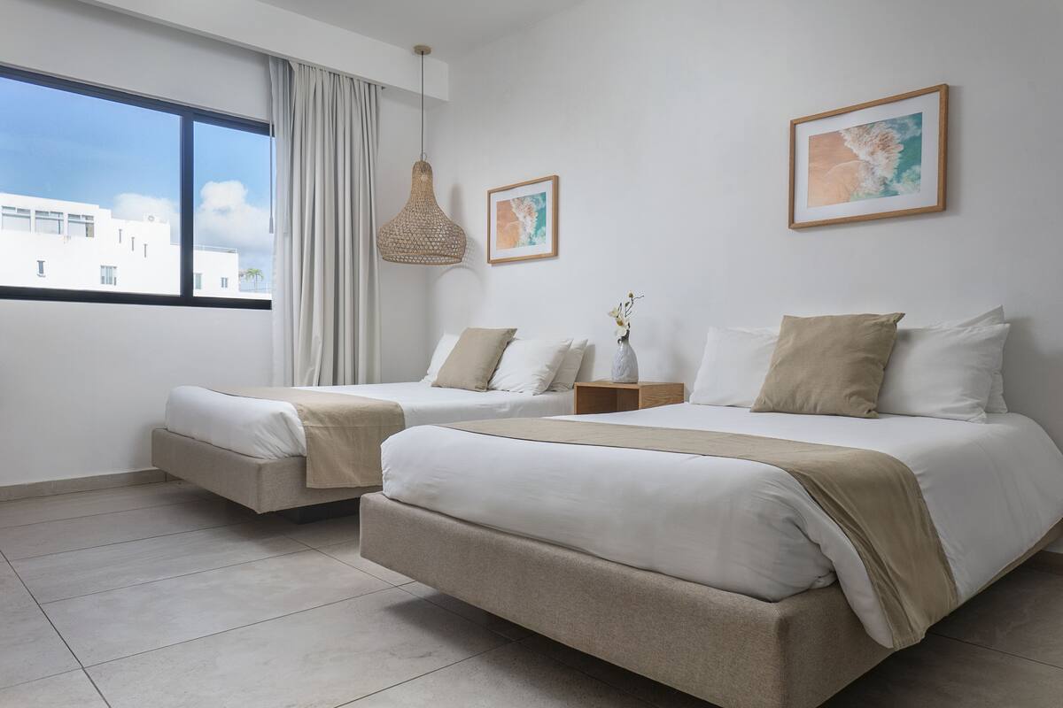 A hotel room with two beds, a large window, a hanging light fixture, and two framed pictures on the wall.