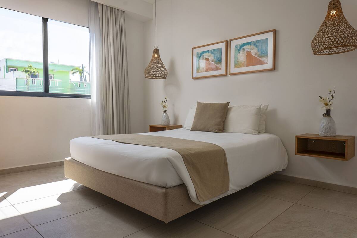 A modern bedroom with a large bed, two bedside tables, a window with a view, and two framed artworks on the wall.