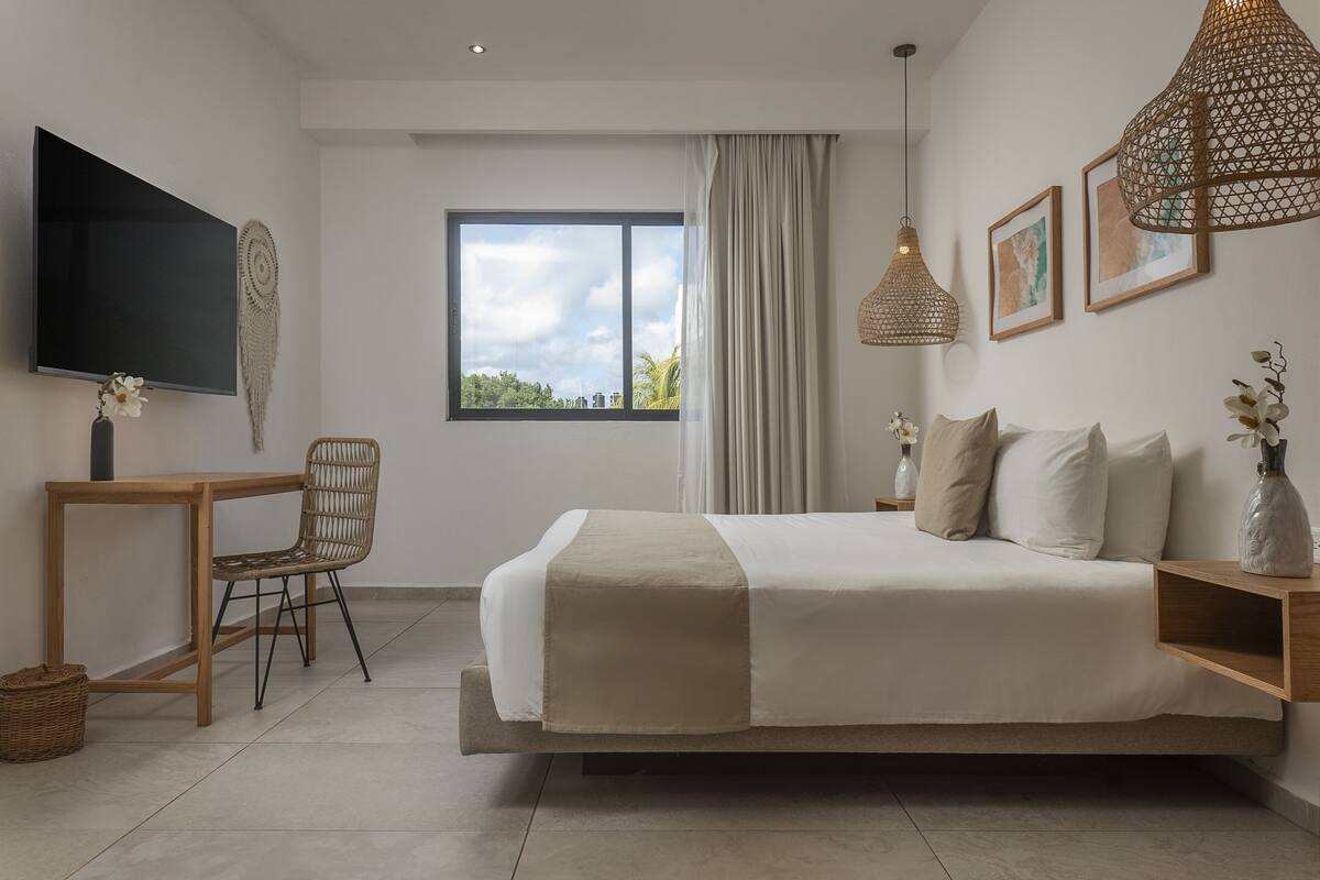 A modern bedroom with a large bed, a TV, a desk, and a window with a view of the city.