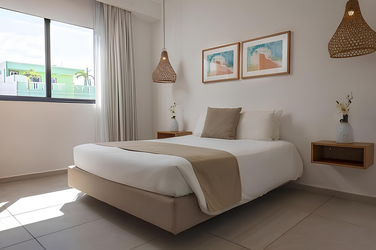 A modern hotel room with a large bed, a view of the outdoors, and two framed pictures on the wall.