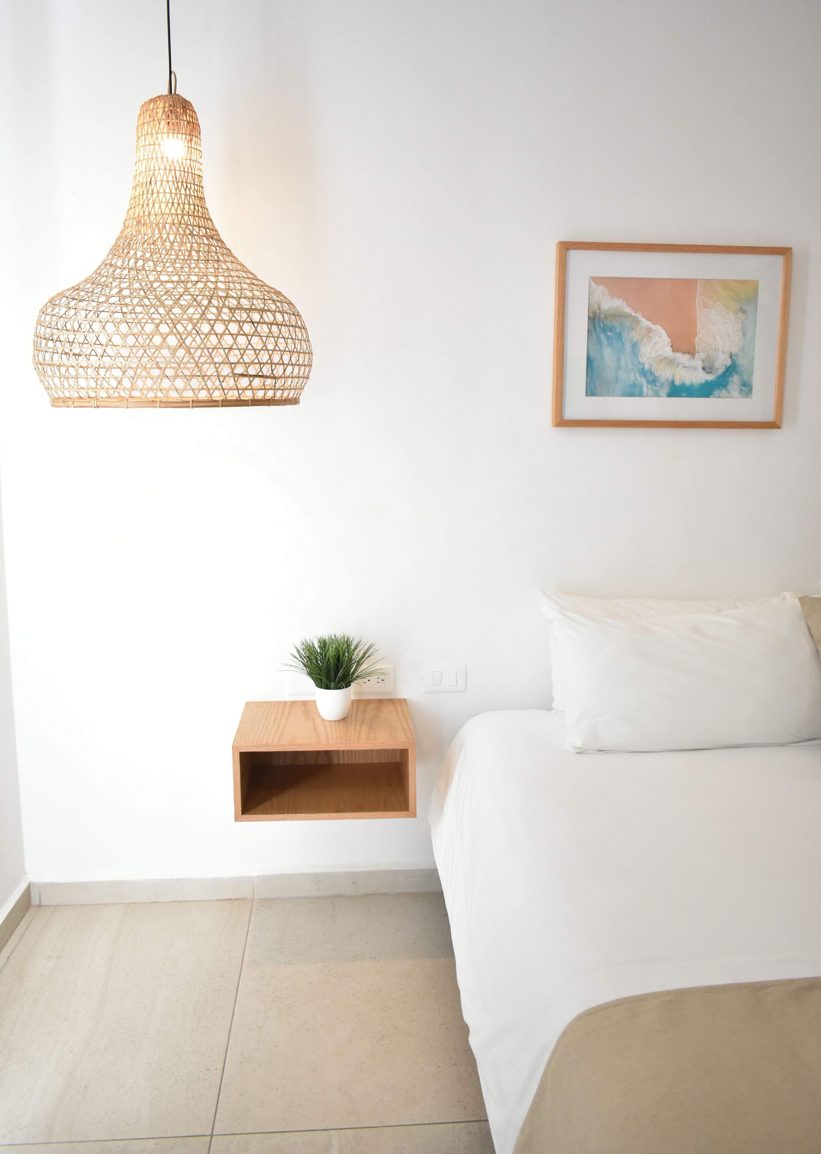 Un dormitorio con una cama, una lámpara colgante, un estante pequeño con una planta y un cuadro en la pared.