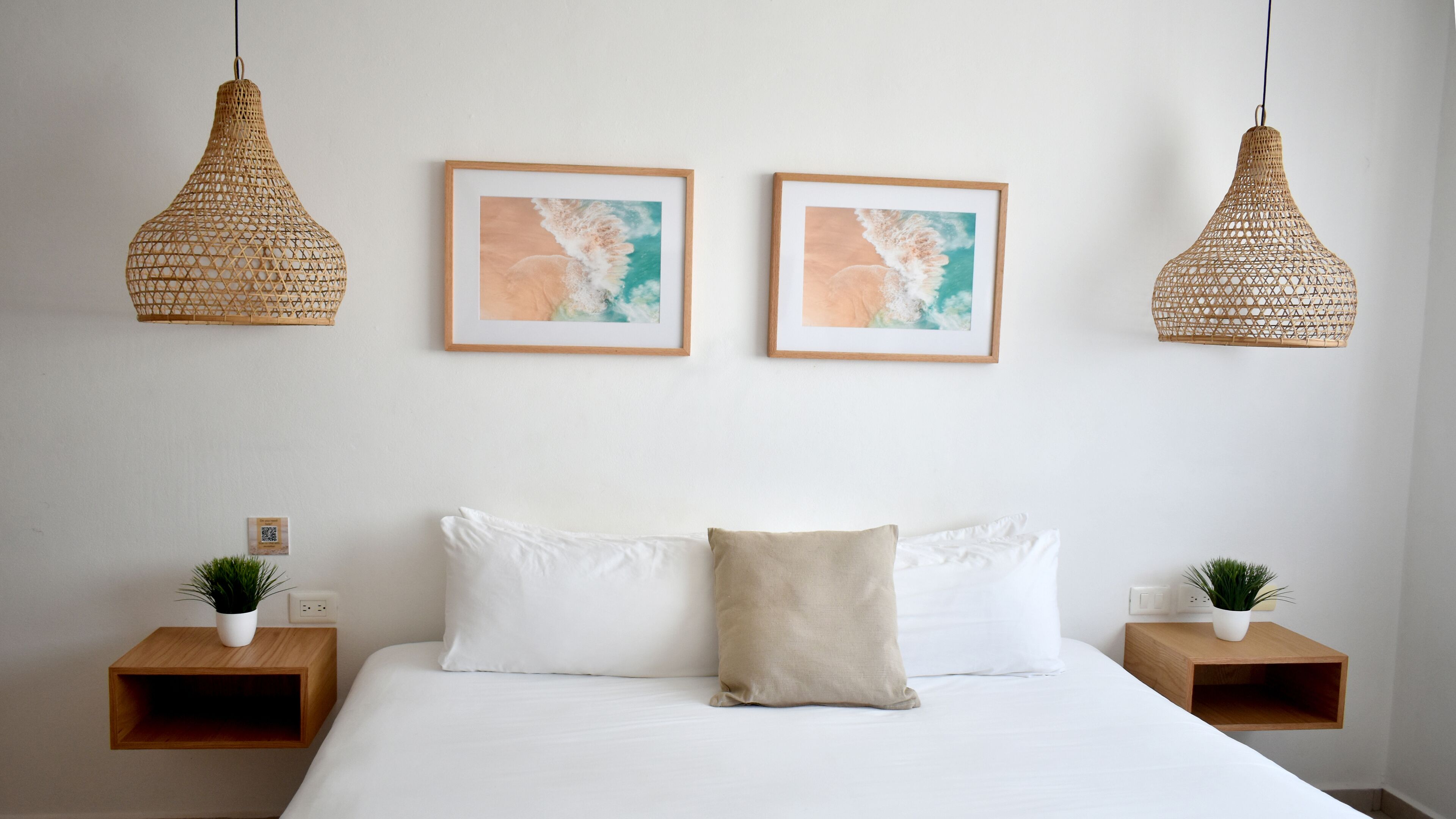 A bedroom with a large bed, two bedside tables with plants, two hanging wicker lamps, and two framed abstract paintings on the wall.