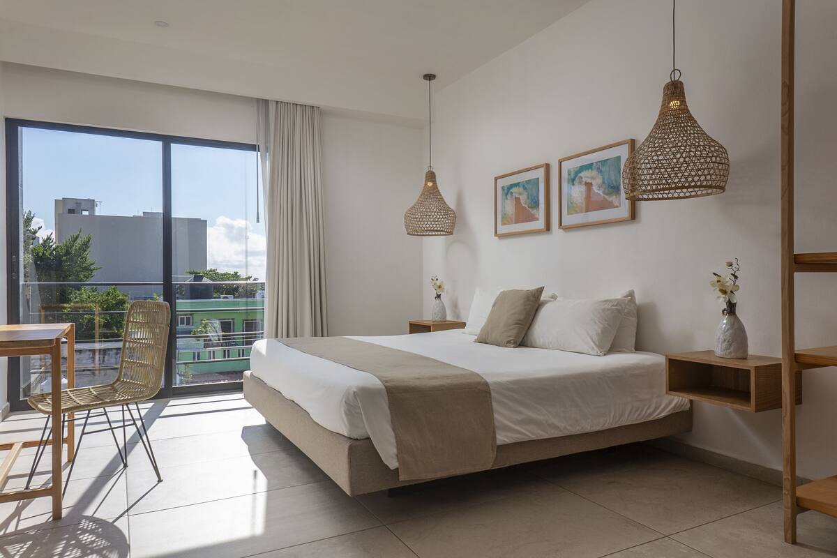 A modern bedroom with a large bed, two pendant lights, a view of the city, and a balcony with a chair.