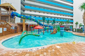 A swimming pool with a water slide, palm trees, and a building with blue and white stripes in the background.