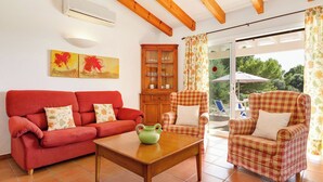 A living room with a red sofa, a wooden table, and checkered armchairs. There is a cabinet with glass doors and a painting on the wall. - Villas Menorca Sur (Alayor)