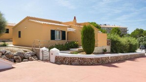A yellow building with a tiled roof, a stone wall, and a small garden with a tall shrub. - Villas Menorca Sur (Alayor)
